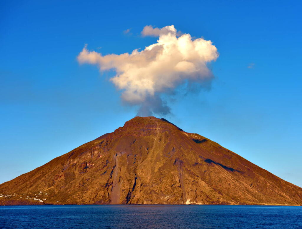 Stromboli Volcano, part of the Aeolian Islands, with plumes of smoke rising from its peak against a clear blue sky.