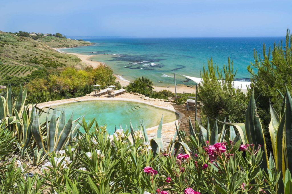  Overlooking a pristine Sicilian beach, this holiday rental villa showcases a beautiful pool amidst lush flora, epitomizing the best of holiday rentals in Sicily.