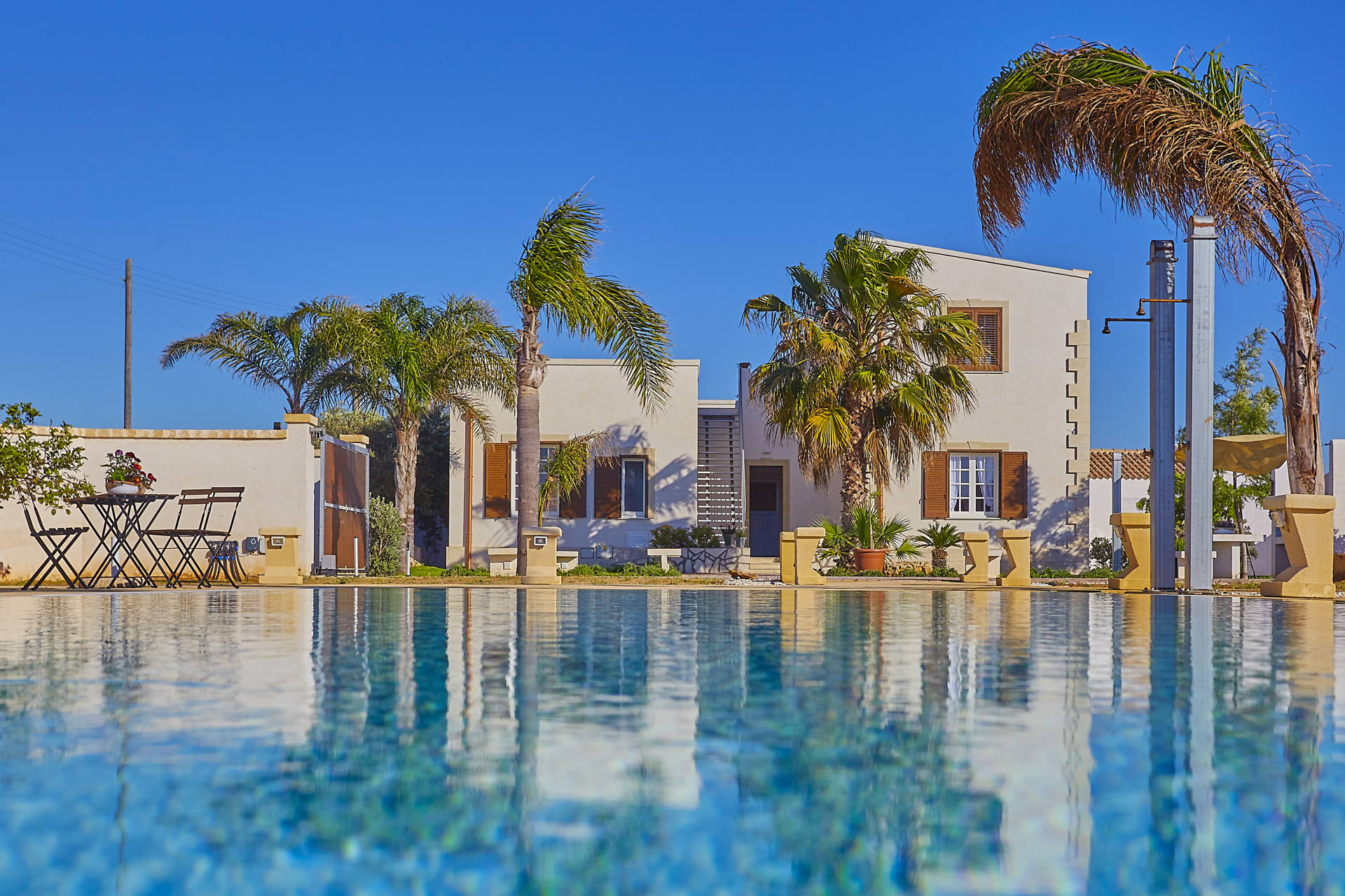 An elegant Sicilian villa with white walls and stone details, surrounded by palm trees and overlooking a clear blue pool under Sicily's serene sky.