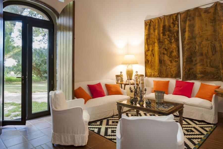 Stylish living room with white sofas, bold cushions, antique wall tapestry, and garden view at Villa Selvira, one of the top-rated Sicily villas with pool.