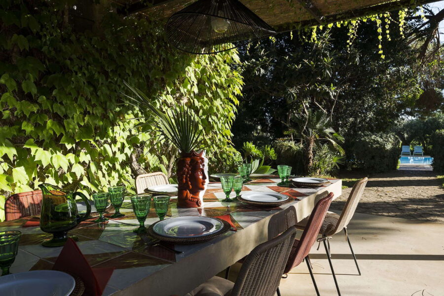 Covered outdoor dining table with ceramic tiles and green glassware, set beneath a vine-covered pergola overlooking the pool at Villa Selvira, Sicily villa with pool.