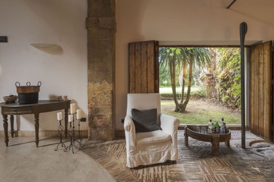 Rustic indoor seating area with white armchair and garden view at Villa Selvira, Sicily luxury villa.