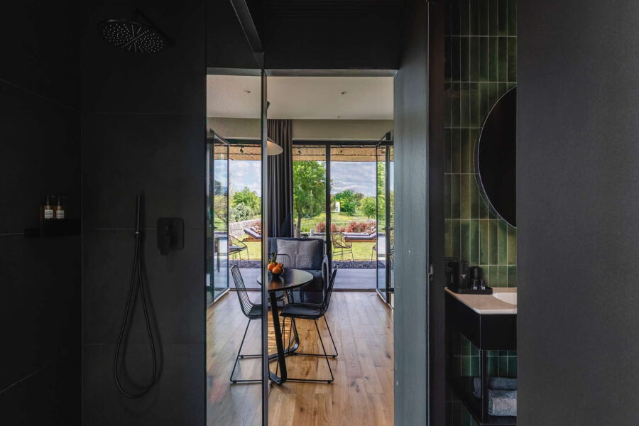 Stylish bathroom with black fixtures and green tiled walls, opening to a bright living area and lush garden at Nature Luxe Cottage in Sicily.